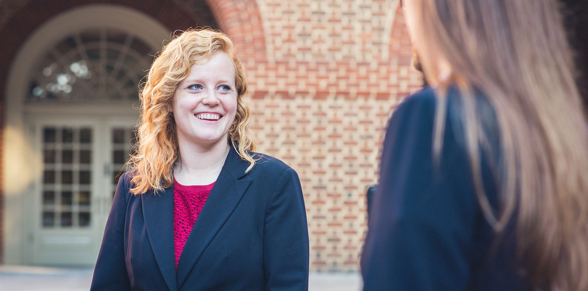 A smiling woman in a navy blazer talks with another person outside a brick building.