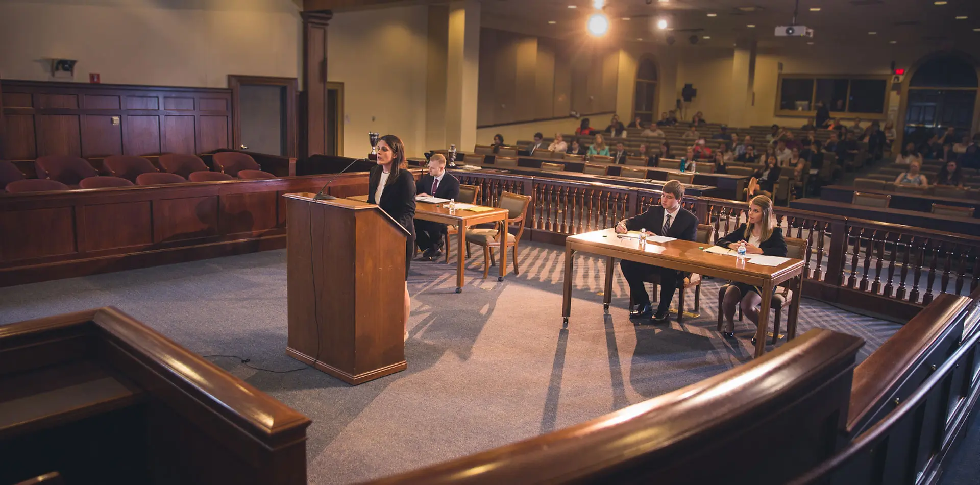 A woman stands at a courtroom podium presenting a case, while suited peers listen attentively and take notes. The setting includes a judge's bench, jury box, and an audience, simulating a real trial environment.