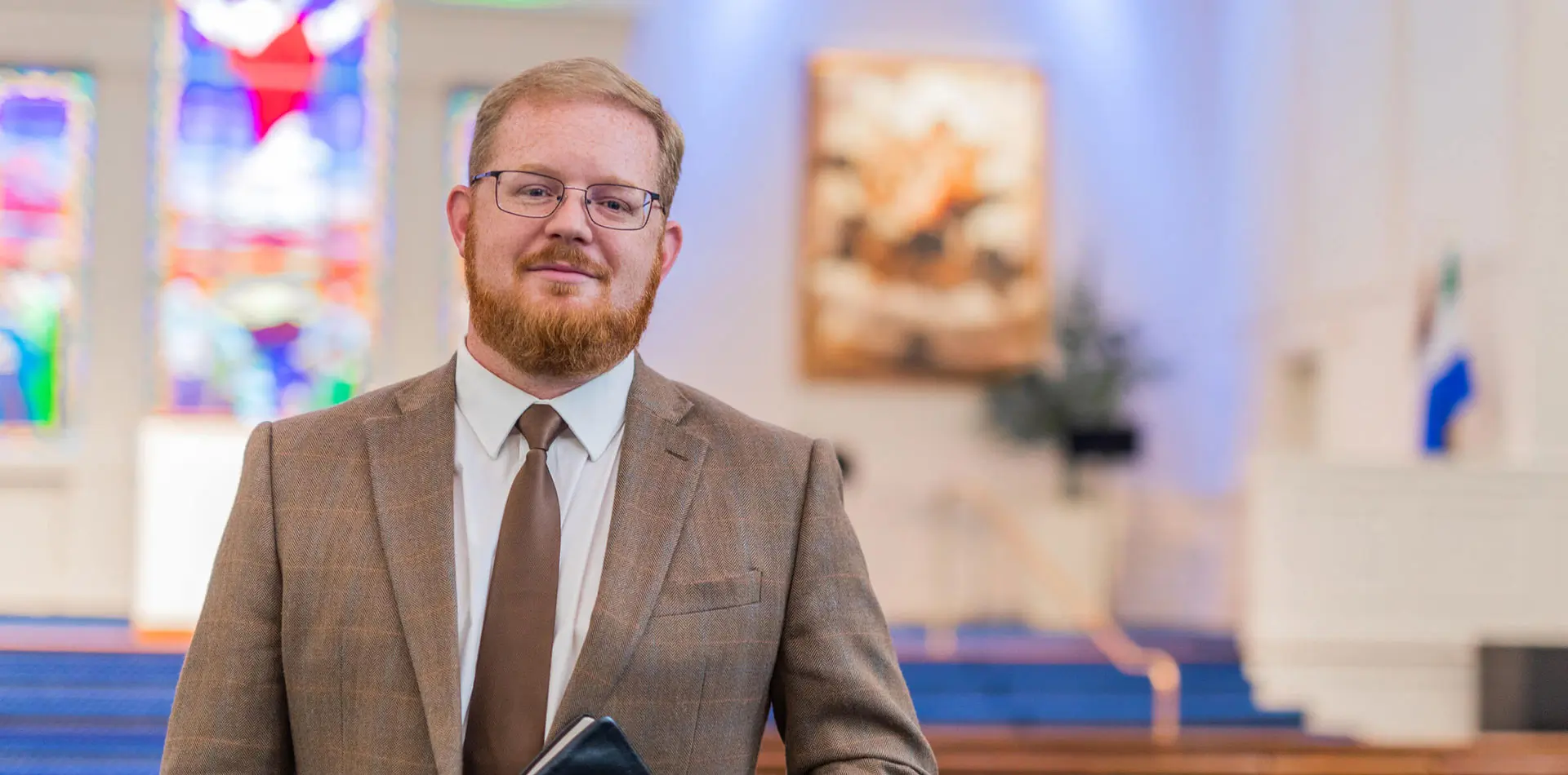 A well-dressed man stands inside a church, holding a Bible and smiling confidently. The background features stained glass windows and soft lighting.