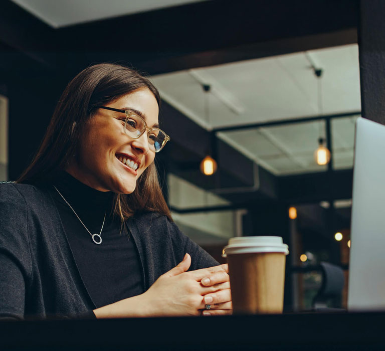 A smiling woman in glasses and dark clothing is seated at a table, looking at a laptop with a coffee cup and lamp beside it.