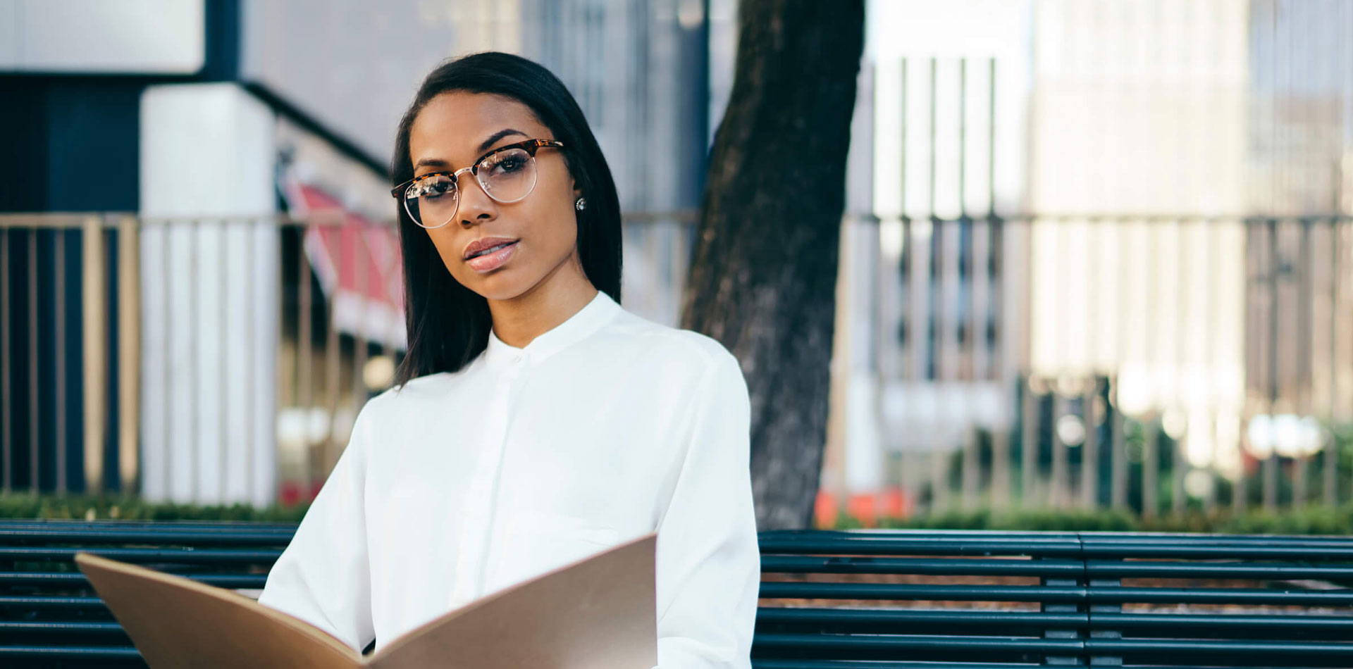 A woman in glasses and a white blouse reads a folder while seated on a bench outdoors.