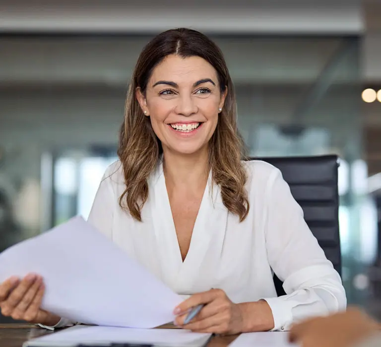 ChatGPT said: A smiling woman in a white blouse holds papers while sitting in a modern office setting.