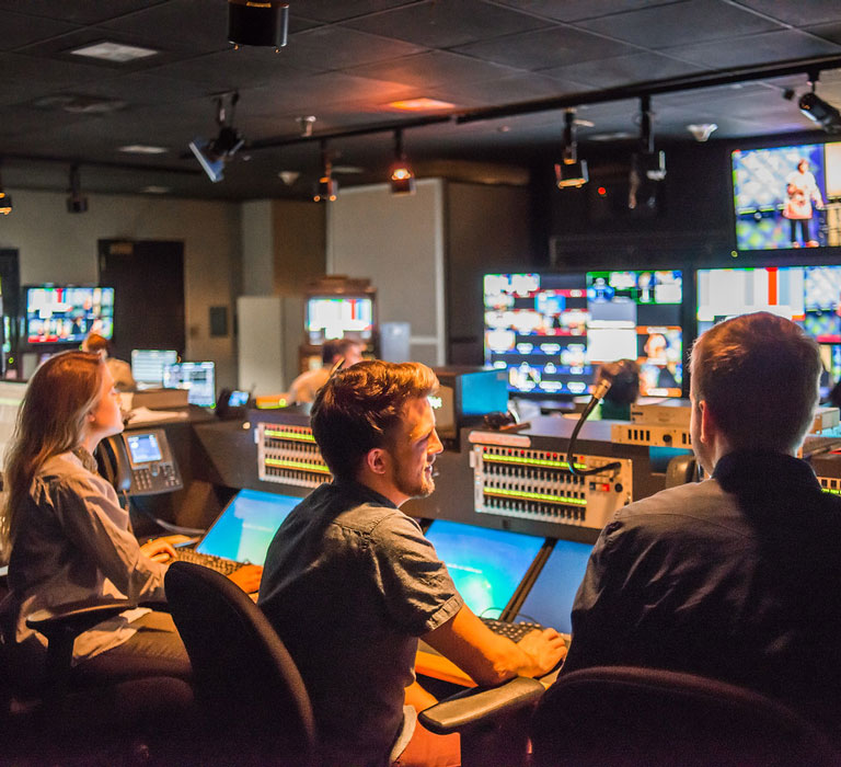 A group of people working in a dimly lit TV control room with multiple screens and equipment.